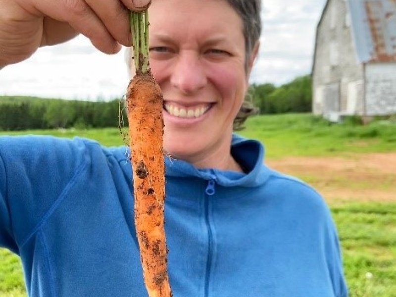 Soleil Hutchinson holds a freshly-dug carrot while standing in a field with barn in background