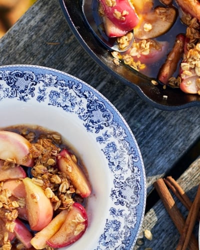 Bowl of apple crisp with cast iron skillet pictured at top of image