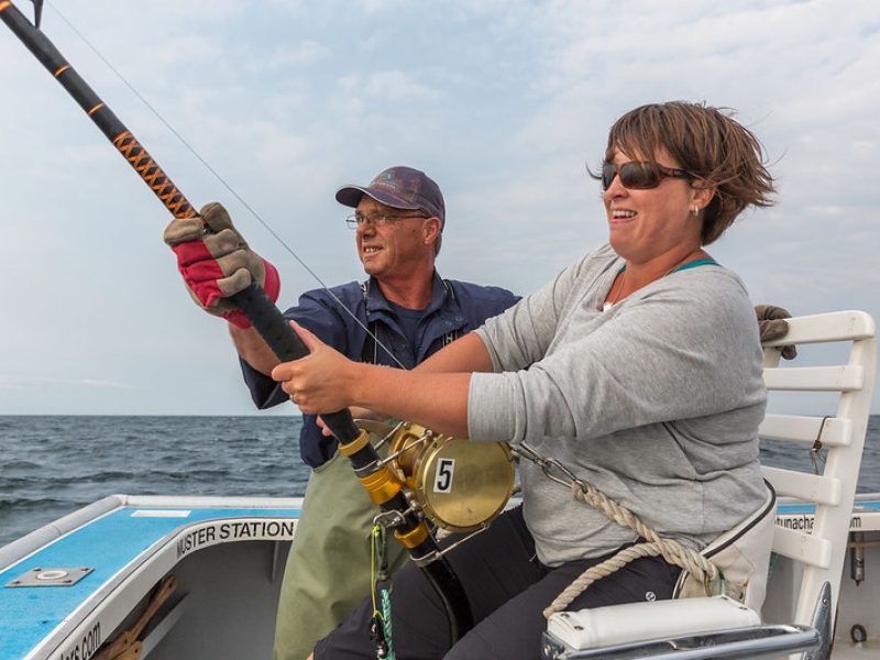 Woman holds tuna fishing rod on charter boat and is assisted by fisher 