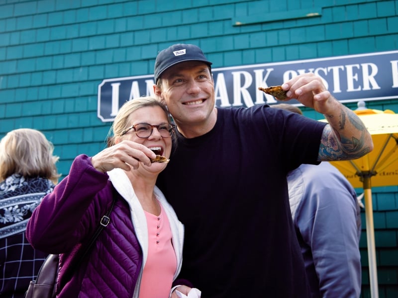 Guest and celebrity chef hold up fresh PEI oysters on half shell in front of marine blue building with "Landmark Oyster House" sign in background