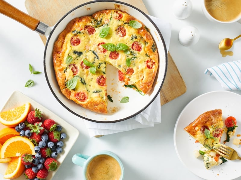 Overhead image of frittata in frying pan and piece served on plate with side plate of fresh fruit