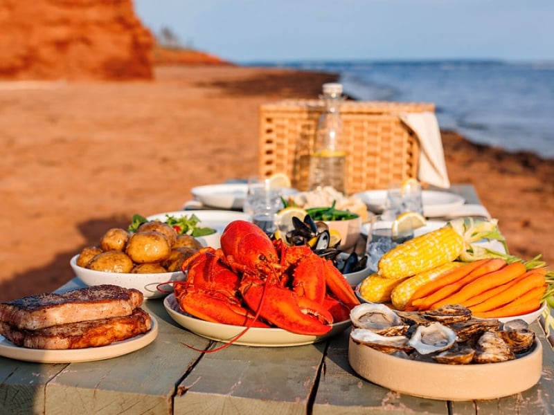 A feast of PEI food product displayed on a picnic table at Cabot Park beach with sand and red cliffs in background