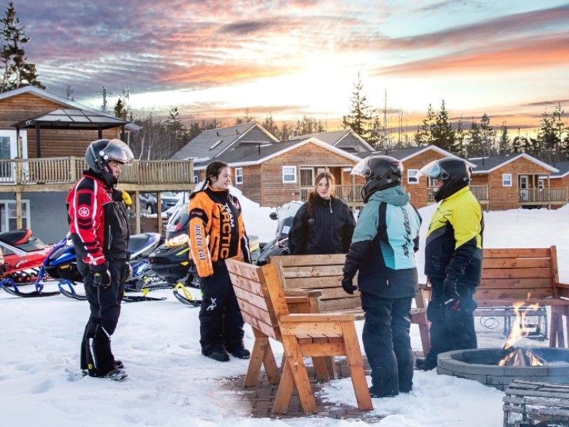 Group of five snowmobilers stand next to outdoor campfire at sunset with sleds and cabins in background