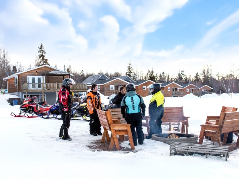 Group of five stand near firepit with snowmobiles and cabins in background