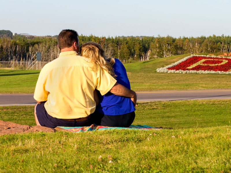 Couple sitting on grass in Borden-Carleton with PEI flower bed in distance