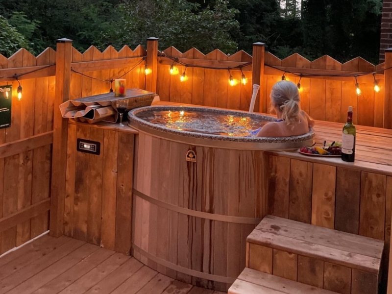 Female sitting in an outdoor cedar hot-tub surrounded by a wooden fence and outdoor lighting 