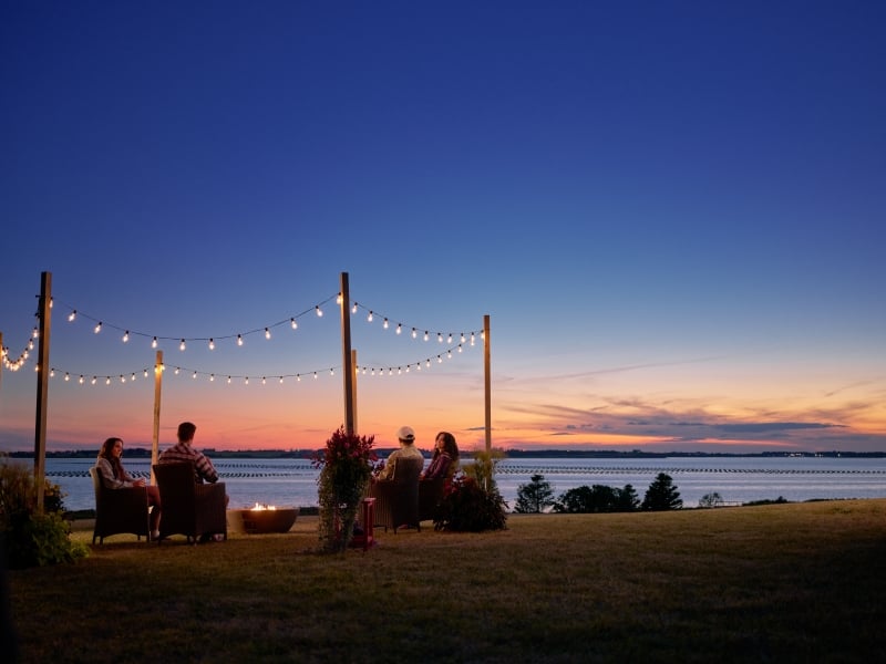 Group enjoys bonfire under patio lights at sunset overlooking the water
