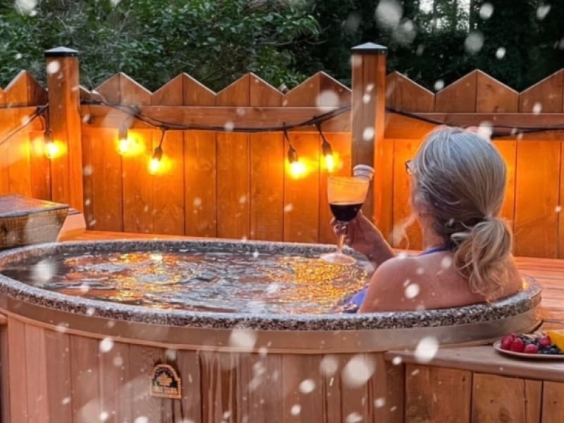 Person enjoys glass of wine in outdoor cedar hot tub as snow flies