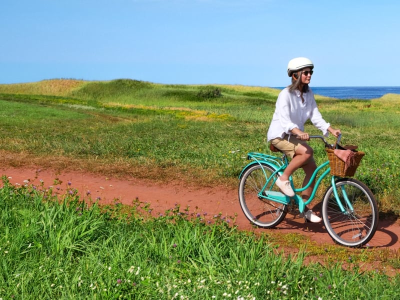 Woman on bicycle on red dirt lane to PEI beach under clear blue sky