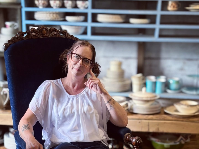 Judith Graves sitting in blue velvet chair in front of selves of antiques