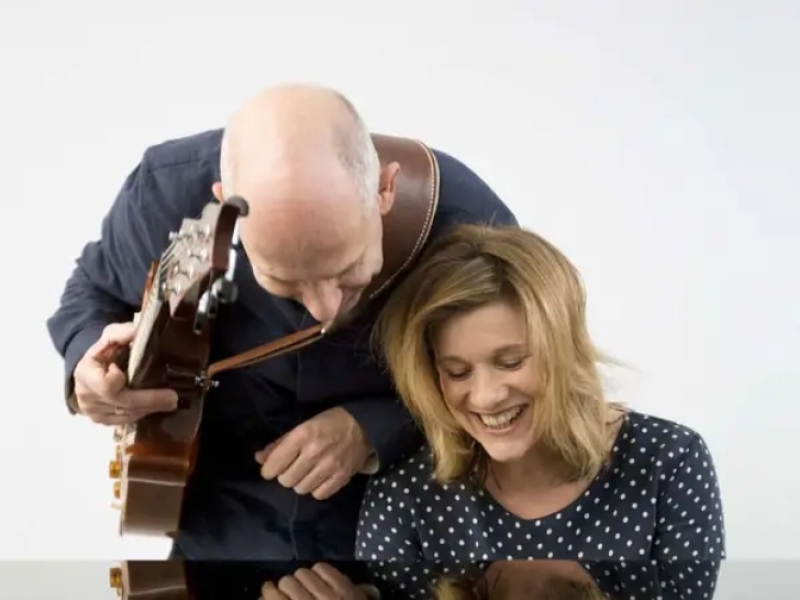 Photo of two musicians one playing piano and the other leaning over them holding a guitar