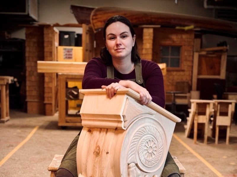 Mel Burke sits behind an elaborate architectural woodwork piece that she made