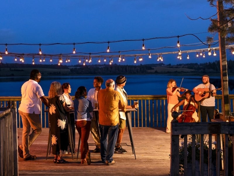 Group watches trio of musicians perform on a riverside deck under patio lights on a summer night