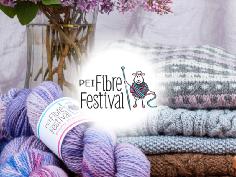 Display of purple yarn skeins, sweaters and a vase of lilacs with PEI Fibre Festival logo in center