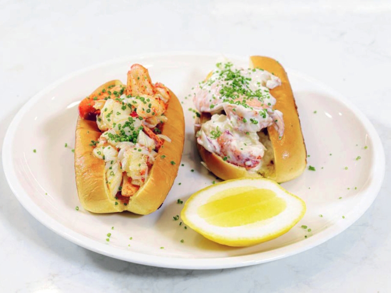Two lobster rolls: One with mayo and one with butter plated with a lemon wedge