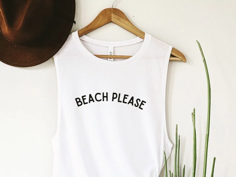 White tank with "Beach Please" screen hanging on white wall with fedora hat beside it