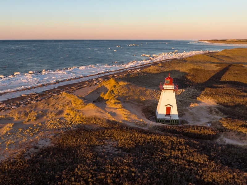 Aerial drone photograph of St. Peters Harbour Lighthouse on Prince Edward Island at golden hour sunset in winter with snow and icy ocean