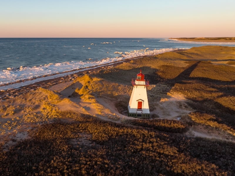 Aerial drone photograph of St. Peters Harbour Lighthouse on Prince Edward Island at golden hour sunset in winter with snow and icy ocean