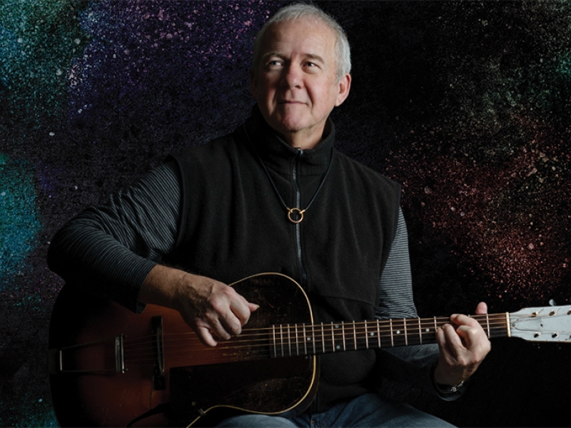 photo of Murray Mclauchlin, sitting playing guitar against a black background, wearing a black vest jeans and grey striped shirt