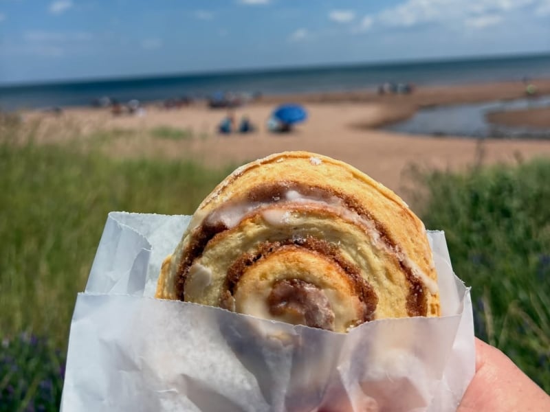 Cinnamon roll in paper sleeve in foreground with beach at PEI Naitonal Park, North Rustico in background