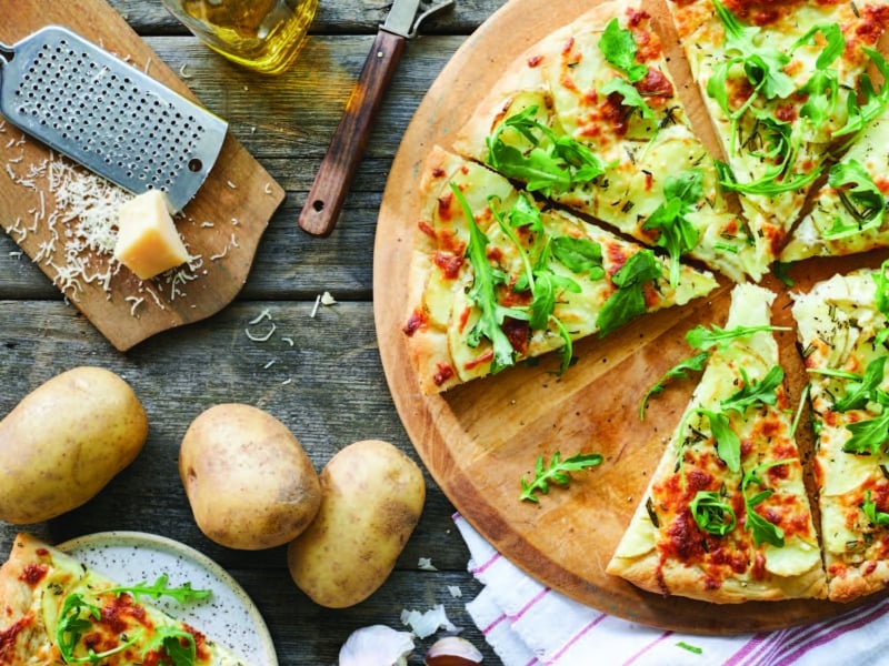 Top view of potato pizza on round cutting board with fresh potatoes and parm cheese to the side