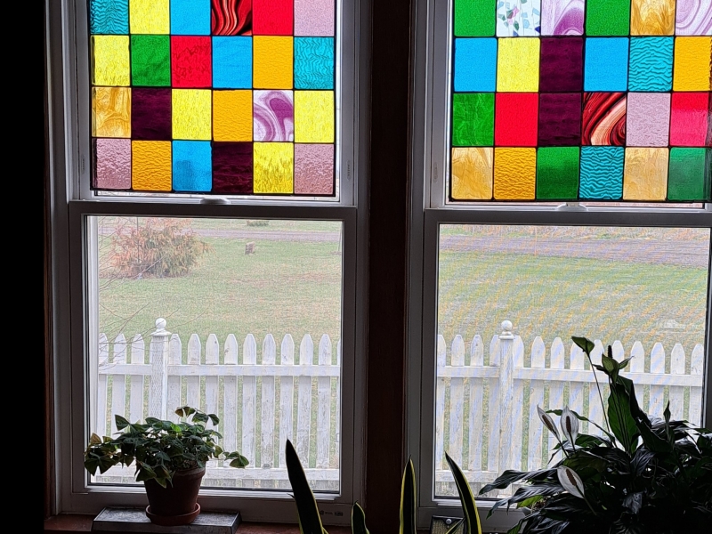 Photo of double windows looking outside. windows have top stain glass panels and plant in front of them