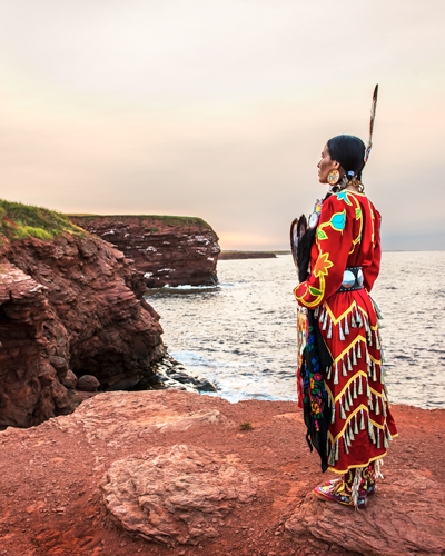 Mi'kmaq woman stands on red rock at Cape Tryon wearing full regalia