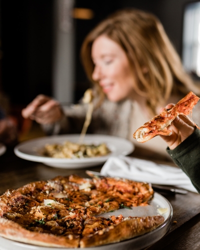 Pizza in foreground with woman eating pasta in background