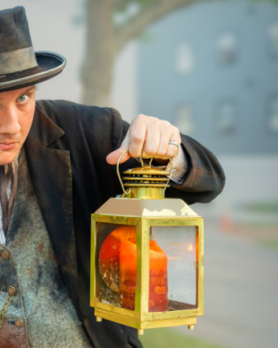 Heritage actor hold lantern with graphic text "Historic Heritage Players Walking Ghost Tour"