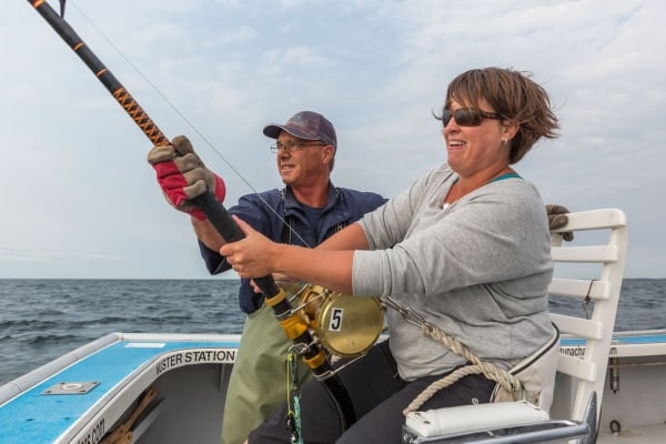 Woman holds tuna fishing rod on charter boat and is assisted by fisher 