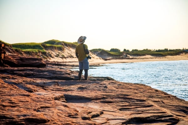 Male holding camera stands on red rock watching the sun set at PEI National Park - Cavendish Beach