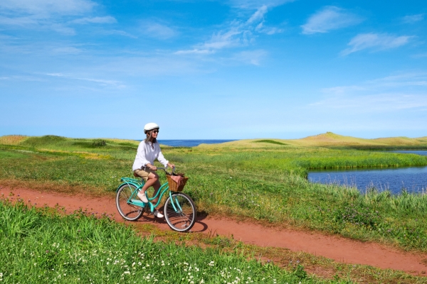 Woman on bicycle on red dirt lane to PEI beach under clear blue sky