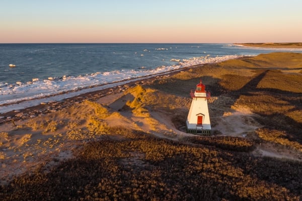 Aerial drone photograph of St. Peters Harbour Lighthouse on Prince Edward Island at golden hour sunset in winter with snow and icy ocean