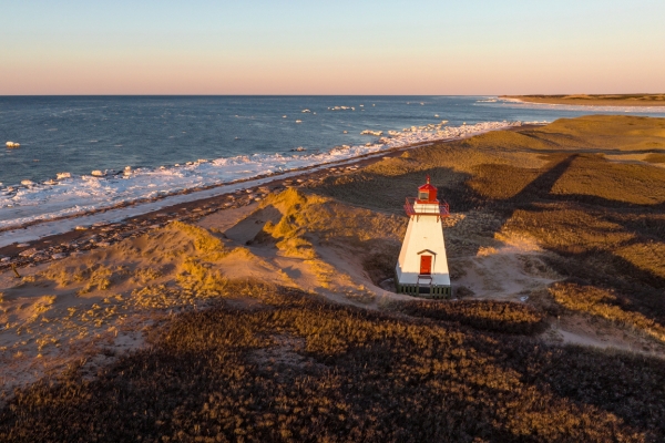 Aerial drone photograph of St. Peters Harbour Lighthouse on Prince Edward Island at golden hour sunset in winter with snow and icy ocean