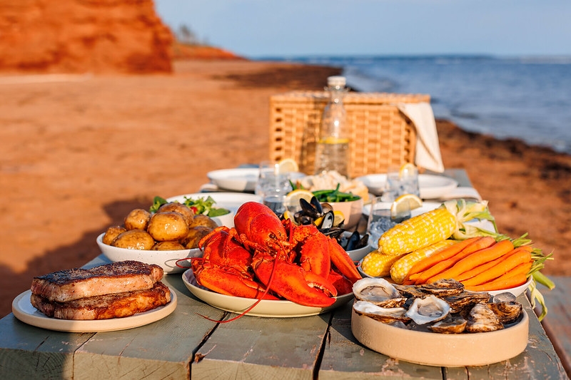 Culinary at Cabot Park A feast of PEI food product displayed on a picnic table at Cabot Park beach with sand and red cliffs in background