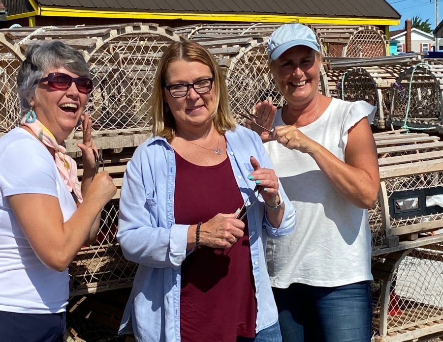 Three women stand in front of lobster traps playing the spoons