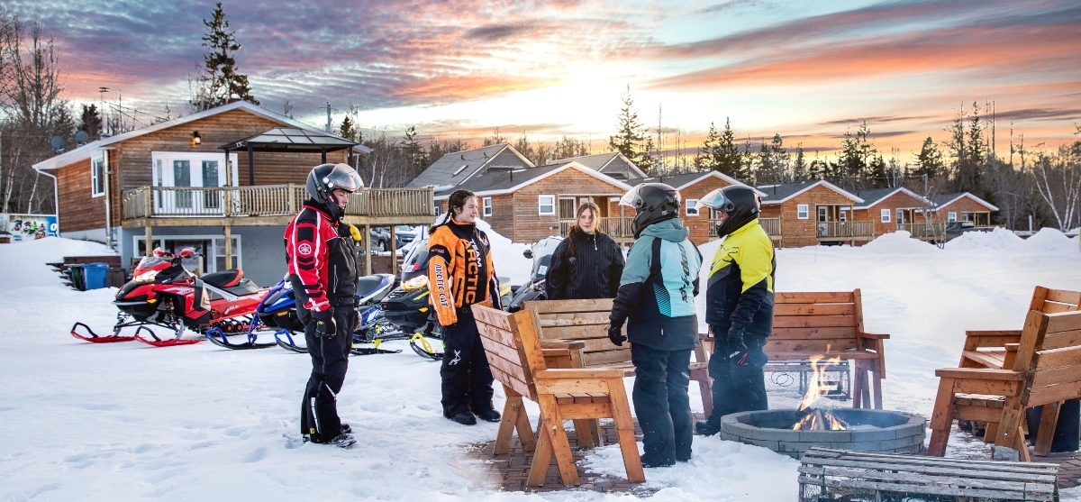 Group of five snowmobilers stand next to outdoor campfire at sunset with sleds and cabins in background