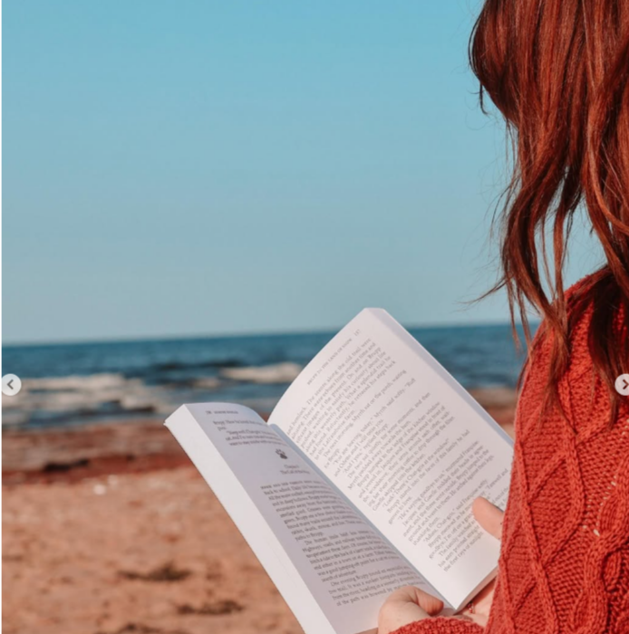 Red haired person sits on PEI beach reading a book