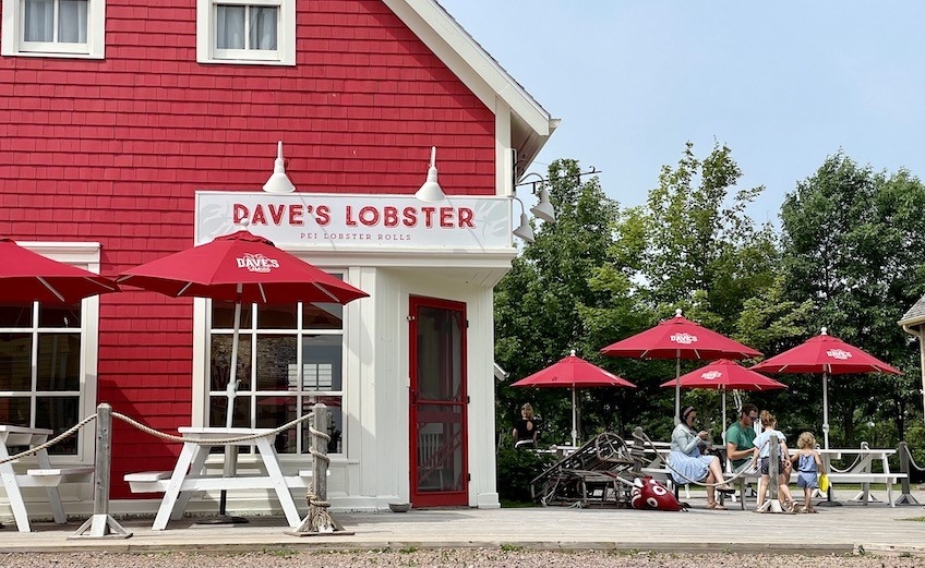 outside view of the corner of the red building which houses Dave's lobster. picnic tables and umbrellas around outside of building