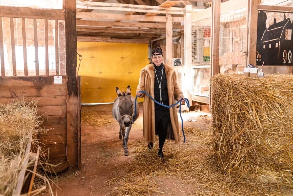 Farmer Flory leads a donkey from stall wearing a fur coat and flapper dress
