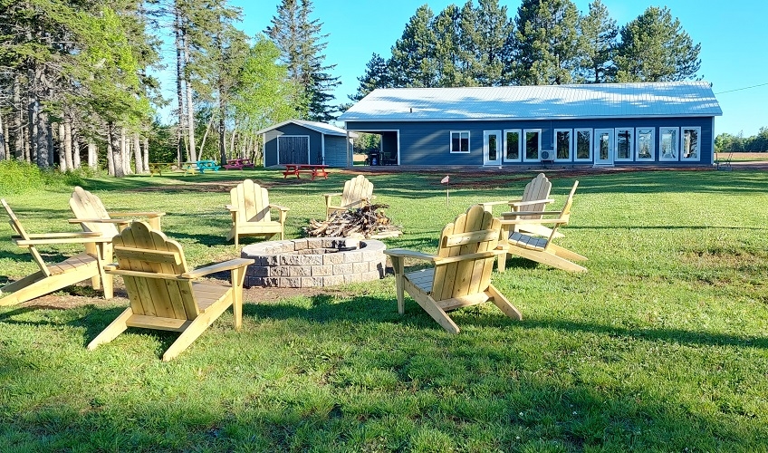 Blue one storey lodge with large grounds, campfire pit circled by Adirondack chairs