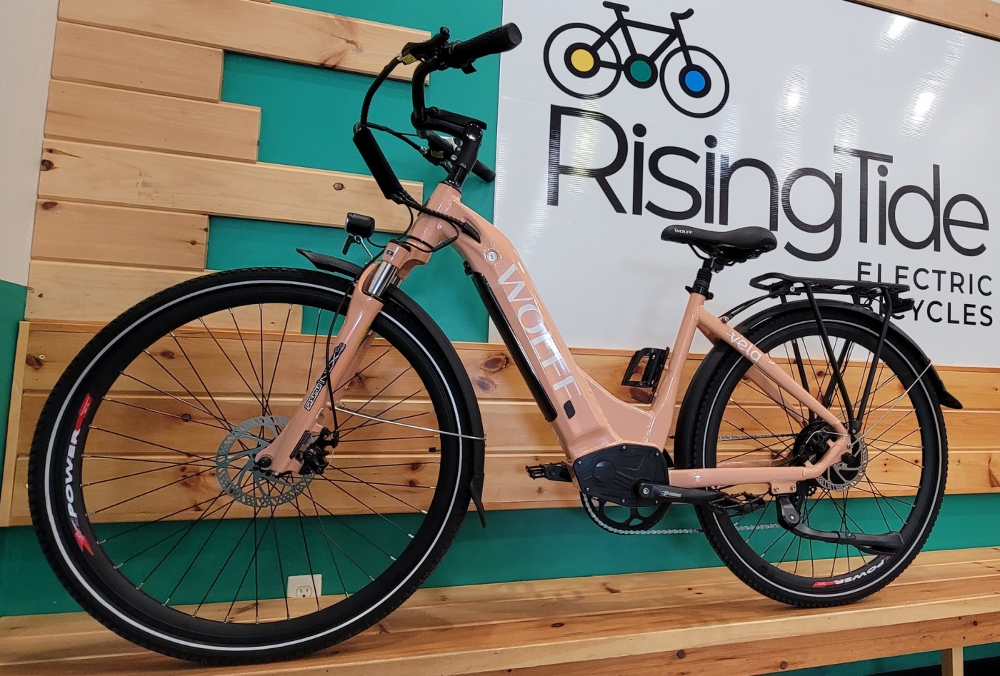 A blush pink electric bike stands in front of a Rising Tide Electric Bikes banner indoors