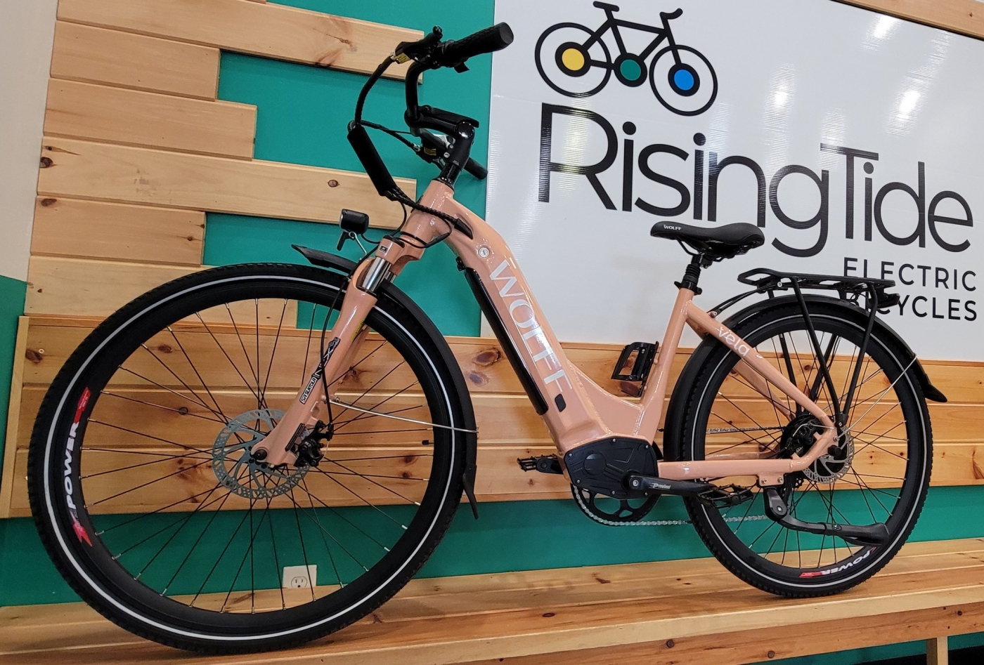 Rising Tide Electric Bike Rentals A blush pink electric bike stands in front of a Rising Tide Electric Bikes banner indoors