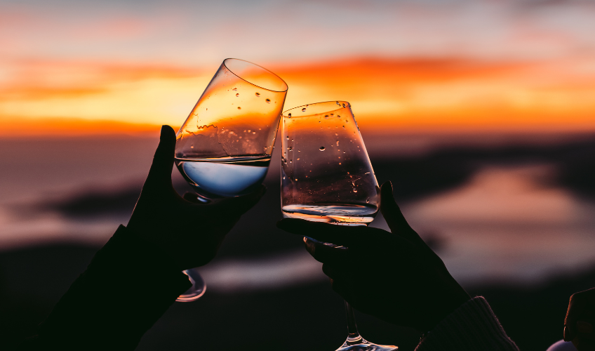 Stock image of couple holding wine glasses together with sunset in background; Source:Canva 