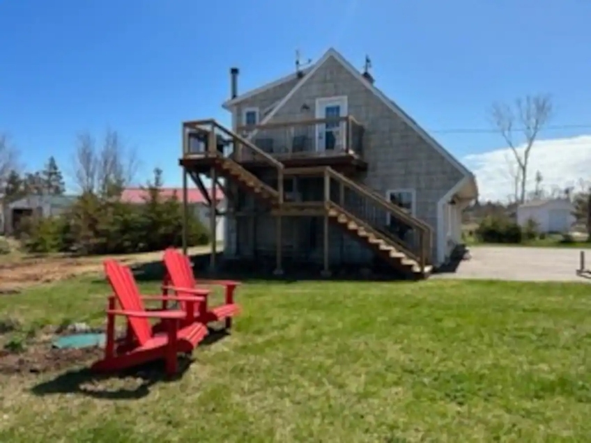 outside view of grey cottage with deck and two red chairs on lawn in front