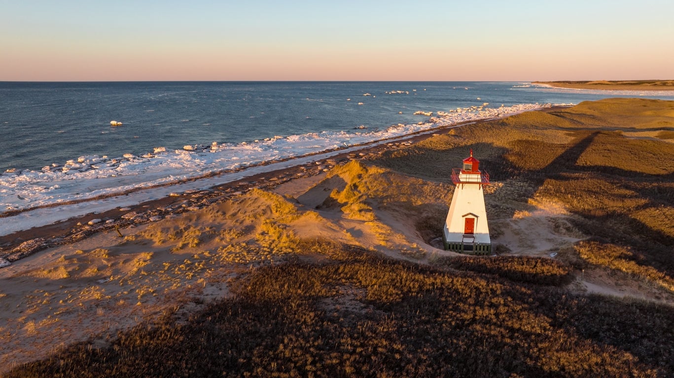 Aerial drone photograph of St. Peters Harbour Lighthouse on Prince Edward Island at golden hour sunset in winter with snow and icy ocean
